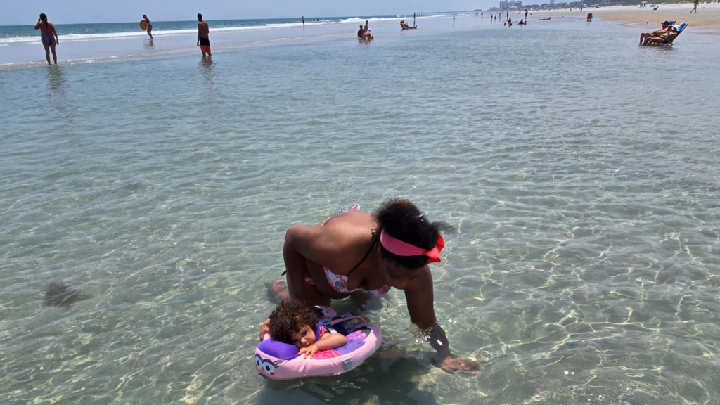 Black mom exploring tide pool with biracial toddler daughter in a floatie on a sunny day at Wrightsville Beach, NC. 1 of 10 Eco-Friendly Beach Activities To Enjoy Without Harming Nature