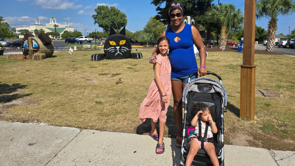 Black Mom and biracial tween daughter and biracial toddler daughter having fun, finding new places in the scavenger hunt. Daughter has peacy skirt and mom has on blue shirt and jeans. Baby is in stroller.